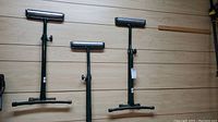 Three green metal roller stands with adjustable height and cylindrical rollers on top, placed side by side on a wood floor.