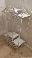White metal lattice frame stand with two removable white wood shelves shown full front and side view on a wood floor.