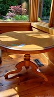 Round pinewood table in natural light near windows, showing tabletop and pedestal base details.