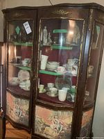 Front view of antique wood china cabinet showing two curved glass doors, ornate gold decorative accents, and painted lower wood panels. Displays porcelain cups and figurines inside.