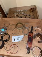 Top-down photo of assorted bracelets and bangles in cardboard box showing variety including metal bangles, wooden bangles, beaded bracelets, leather cuff, charm bracelets