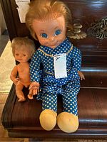 Front view of two dolls sitting side by side on a wooden surface, Mrs Beasley doll in blue polka dot dress, smaller anatomically correct male doll nude with jointed limbs.