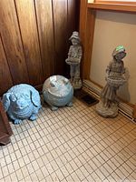 Four garden ornaments including the boy figure holding a platter, the girl figure holding a basket, a frog, and a dog, all placed indoors on a tile floor against wood-paneled walls. All are blue-grey with weathered appearance.