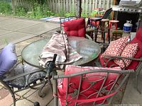Photo of the round outdoor table with glass-like top, metal frame chairs with red and patterned cushions around it, also includes some pillows and an umbrella folded on the table