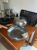 Photo showing assorted pots including stack of Revere Ware pots with lids and cast iron fry pans, Monix fry pan with glass lid, large fry pan, muffin pans, and cookie sheet on a wooden table.