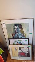 Three framed art prints stacked against a wall. Largest one depicts a cubist portrait style image. Medium sized Picasso print is below. Smallest framed print shows a colorful round natural design.
