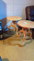 Two matching round wood side tables with different wood grain patterns, placed on a carpeted floor near a dark leather chair and heater.