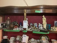 Wide view showing multiple ceramic and porcelain items including a tall alabaster figure, small figurines, vases, and decorative items on two shelves.