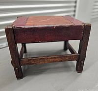Front view of antique wooden foot stool showing wooden frame and leather seat with metal corner accents.