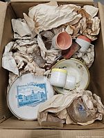 Top view of the box showing various vintage ceramics and glass items wrapped in paper including decorative plates, small cups, and crystal pieces.