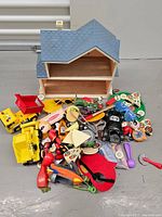 Wooden dollhouse and assortment of toys scattered in front of it including dump trucks, play food, and figurines.