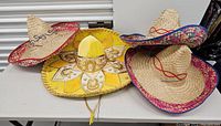 Four Mexican sombreros displayed on a table including one yellow decorated sombrero and three natural straw sombreros with colored trim.