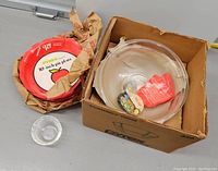 Photo showing three large nested vintage Pyrex clear glass mixing bowls with lids inside a box labeled Pyrex, alongside pie plates wrapped in paper and a small glass bowl.