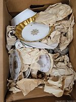 Box showing multiple ceramics wrapped in brown paper including a white scalloped bowl with gold trim and floral center motif, and decorative plates