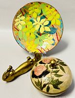 Enamel floral metal plate standing on clear display with brass dog utensil rest and embossed floral covered brass bowl in front