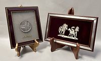 Two framed pewter plaques: one depicting horses signed Q. Pasquini marked 925, and one religious plaque. Both displayed on wooden easels.