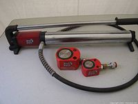 Photo of a silver hydraulic power pack with black end caps labeled 'Big Red', alongside two red short stroke hydraulic pistons, one with a connected hydraulic hose.