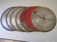 Seven circular saw blades of approx. 12 inch diameter, with 60 to 80 teeth, laid out overlapping on a table surface. Some blades have colored painted sections in blue and red.