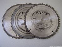 Photo showing three used 10-inch circular saw blades stacked slightly overlapped, branded Hitachi, showing wear and use marks.