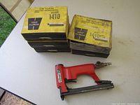 Red Haubold wire upholstery stapler with six yellow and black boxes of Grip Clinch wire staples labeled 1410, stacked on a white table.