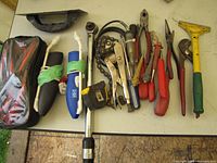 Photo shows an assortment of hand tools laid out on a table, including pliers, wrenches, a tape measure, a putty knife, a hammer, a chain wrench, screwdrivers, and a tool pouch.