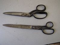 Two large scissors placed side by side on a flat surface. Both have black handles and metal blades.