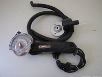 Photo showing the Rotozip electric zipsaw with black hose attachment, an extra circular saw blade, and a protective metal disk laid out on a light-colored surface.