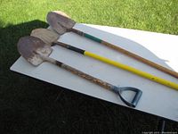 Photo showing three garden shovels laid out on a white table outside on grass. The shovels vary in handle design and paint, with signs of use on the blades and handles.