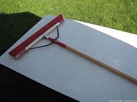 Photo of a 24 inch heavy duty broom lying on a white table outdoors with a background of green grass. The broom has stiff red bristles with a red metal frame and a wooden handle.