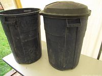 Photo showing two large black Rubbermaid waste bins on a table outdoors, one with a lid and the other without. The bins show dirt, scuffs, and wear.