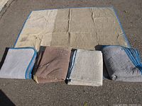 Five folded padded moving blankets in various colors laid out on pavement, one unfolded flat showing size and quilting pattern