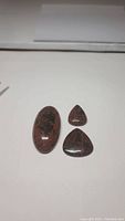Three mahogany jasper cabochon stones on a white surface, showing oval and pear shapes and natural veining pattern