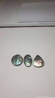 Three natural abalone doublet gemstones displayed on a plain white surface, showing their multi-shape forms and iridescent color patterns
