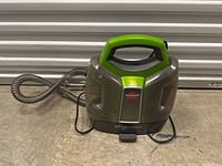 Front view of Bissell Little Green Pro portable carpet cleaner with green handle, clear side tanks, and attached hose, placed on concrete floor.