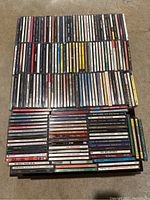 Overhead view of a large set of stacked music CDs in jewel cases showing album spine titles and designs, arranged in rows and columns on a flat surface.