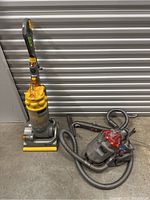 Dyson upright and canister vacuum cleaners shown together on floor in front of a shutter door.