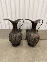 Pair of dark metal pitchers with floral embossed design, curved handles, and flared spouts on floor against white background.