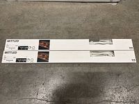 Two boxed IKEA MITTLED LED lights laying on concrete floor, showing full packaging and partial cords through box windows.