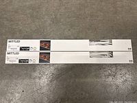 Two IKEA MITTLED boxed LED under cabinet light fixtures shown side by side on floor, unopened in white packaging with product info and photo of light in use.