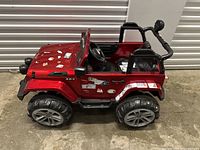 Side view of red kids electric Jeep with stickers, two seats, and large wheels.