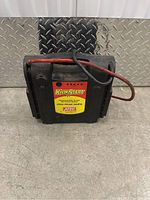 Front view of black rectangular KwikStart power supply with red and black cables wrapped over handle, placed on concrete floor with metal background.