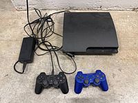 Top view of the PS3 Slim console, two wireless controllers (black and blue), and power adapter with cables spread out on a surface.