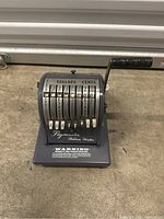 Front angle view of gray Paymaster Ribbon Writer check writer showing dollar and cent dials and crank handle.