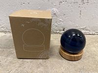 Blue decorative sphere with dark and light swirled patterns sitting on a smooth wooden base next to its cardboard box, set against a concrete floor and white textured background.