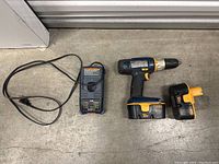 Photo showing Ryobi cordless drill, spare 18V One+ battery, and battery charger with power cord laid out on concrete floor