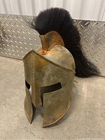 Front angle of a metal Spartan style helmet with black horsehair crest on a gray floor with metal diamond plate background.