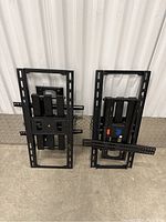 Two black TV wall mount brackets standing vertically against a white corrugated wall on a concrete floor. Both have rectangular frames with multiple mounting slots and horizontal bars.