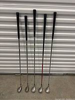 Six vintage Taylormade Burners golf clubs standing upright, showing grips and shafts with one red shaft among mostly silver shafts.