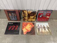 Six vinyl record albums arranged on concrete floor, showing front cover art: Emmylou Harris, Van Morrison, The Police, Eagles, The Doors, and one with a red cover featuring a close-up of a person singing (likely Jimi Hendrix).