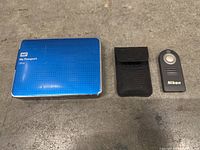 Front view of blue WD My Passport Ultra external hard drive, Nikon remote with pouch on concrete floor.